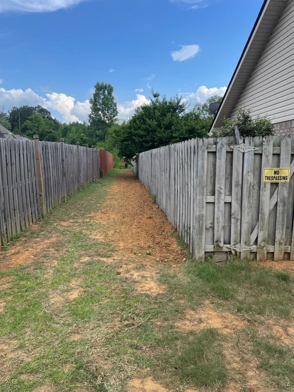 Narrow dirt path between weathered wooden fences, leading toward trees and a house.