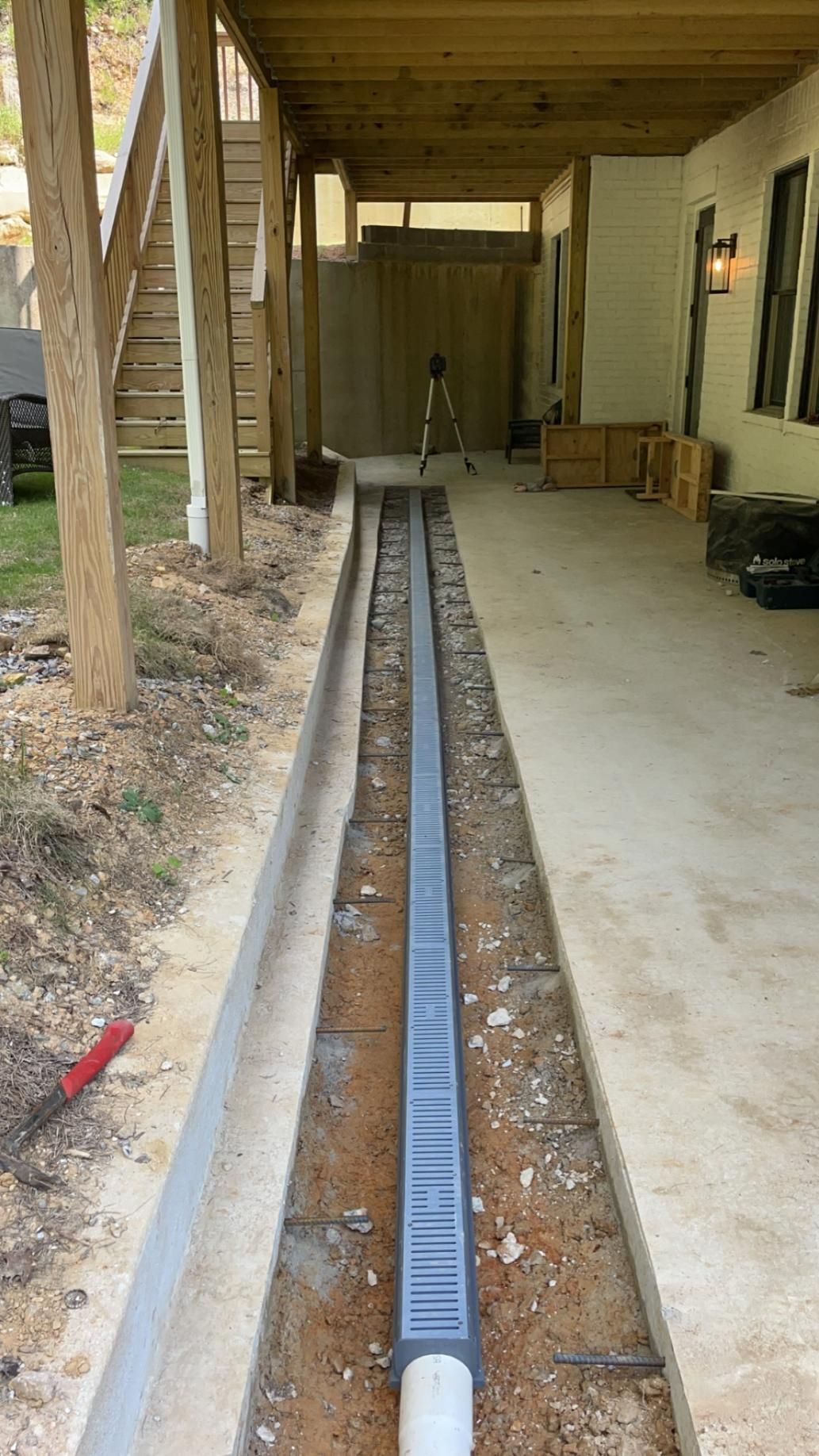 A narrow trench with a grated drain installed on a concrete path under a wooden deck.