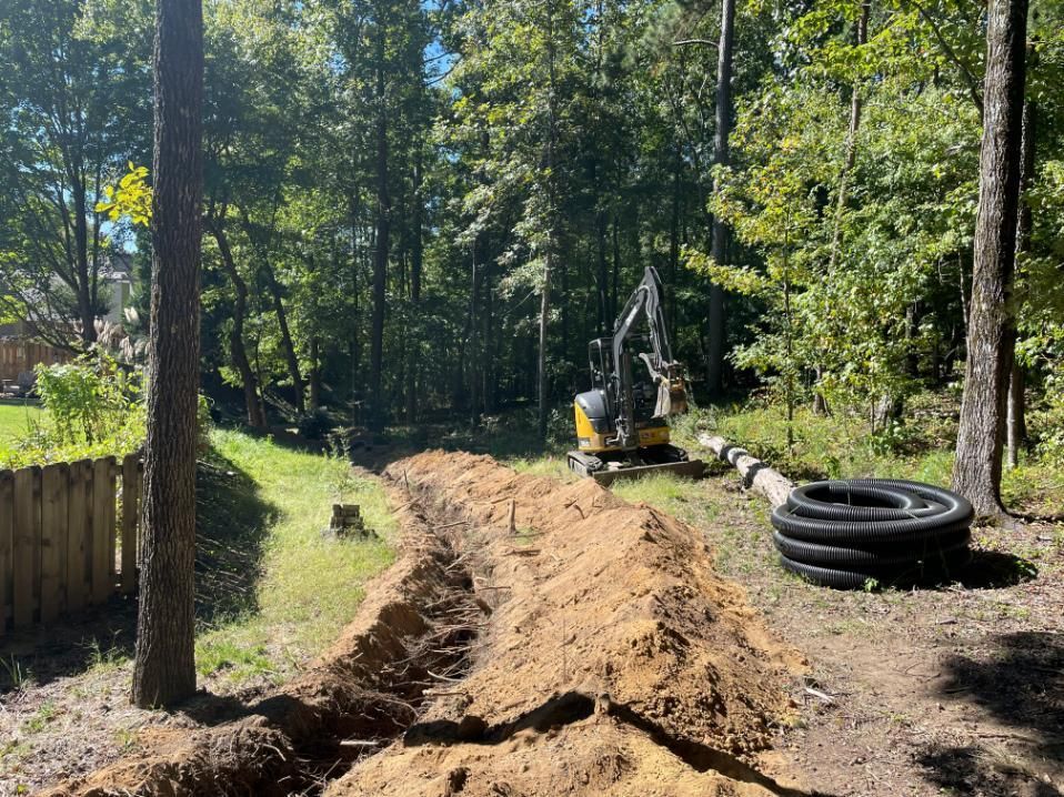 A trench being dug in a wooded area with an excavator and drainage pipes.