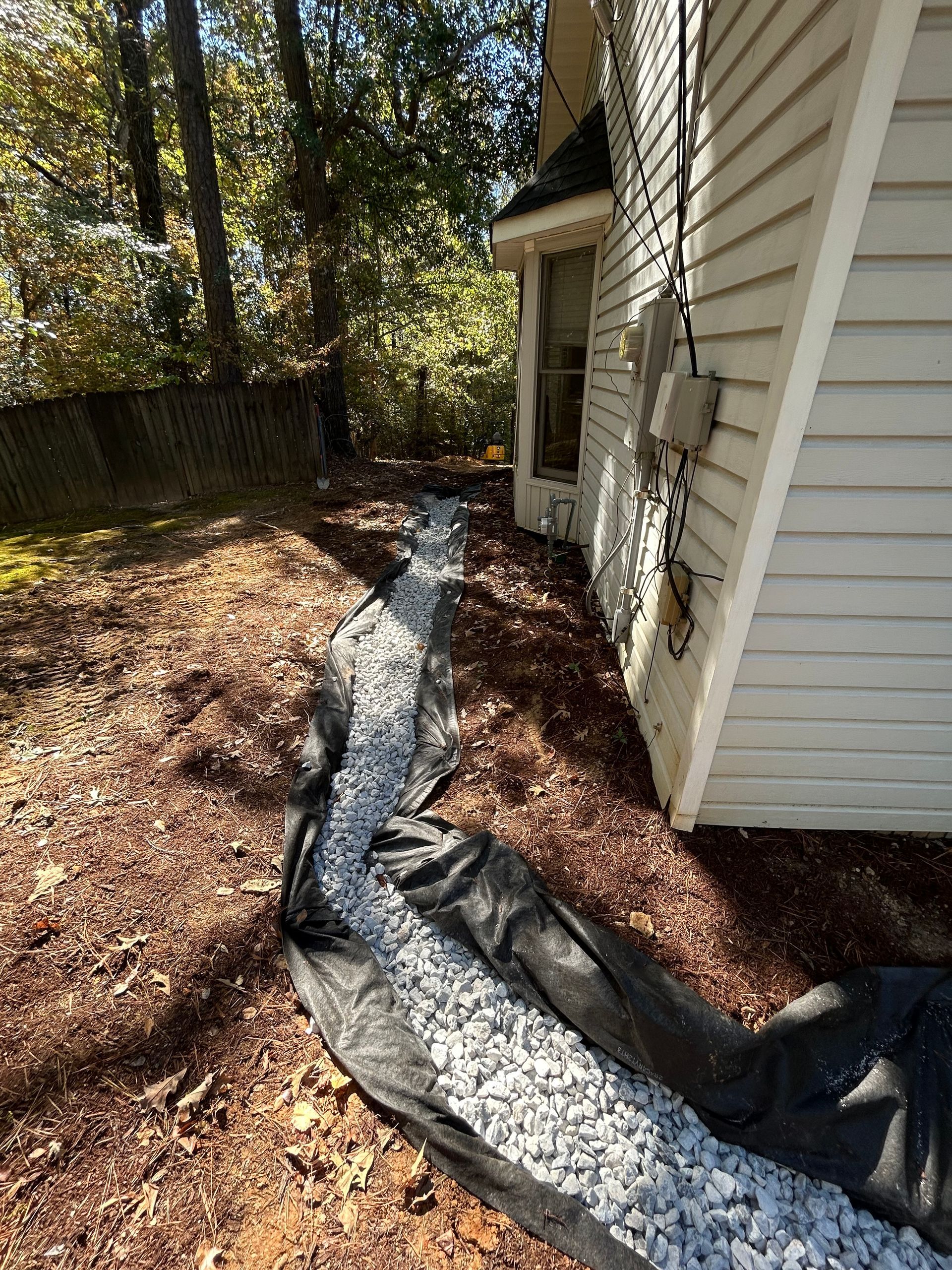 Trench filled with gravel, lined with black fabric, near a white brick house.