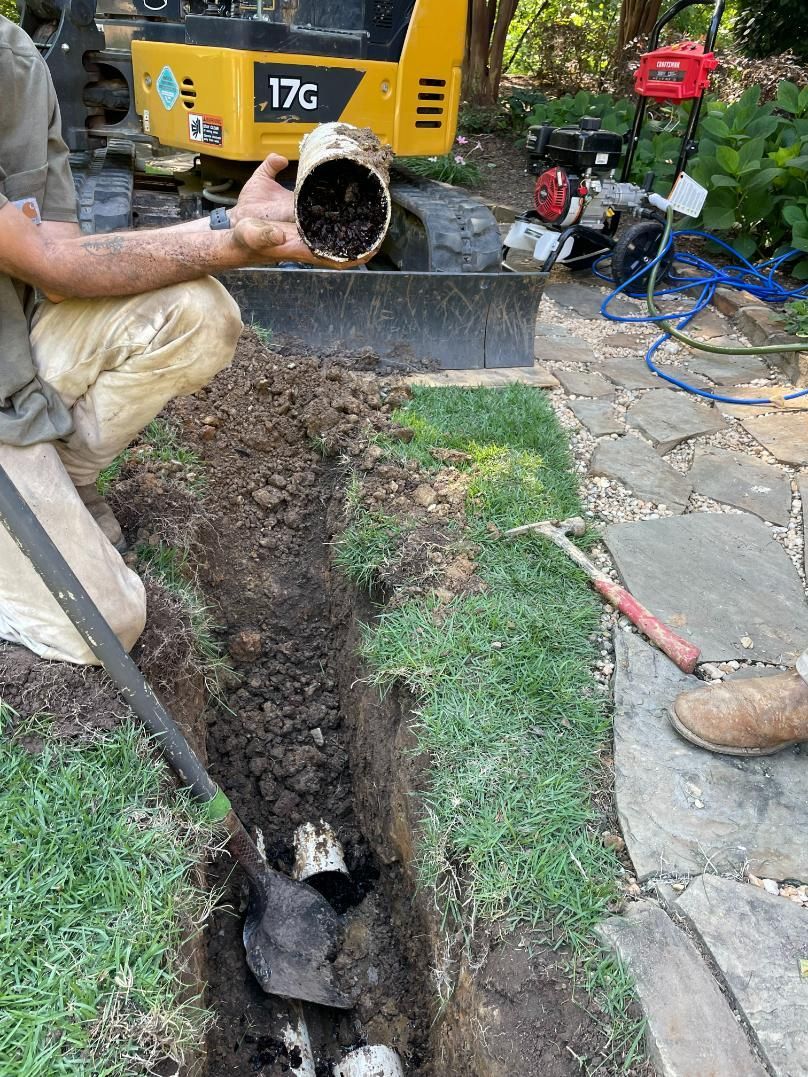 Man in trench holding pipe, working on a plumbing project outdoors. Soil and tools visible.