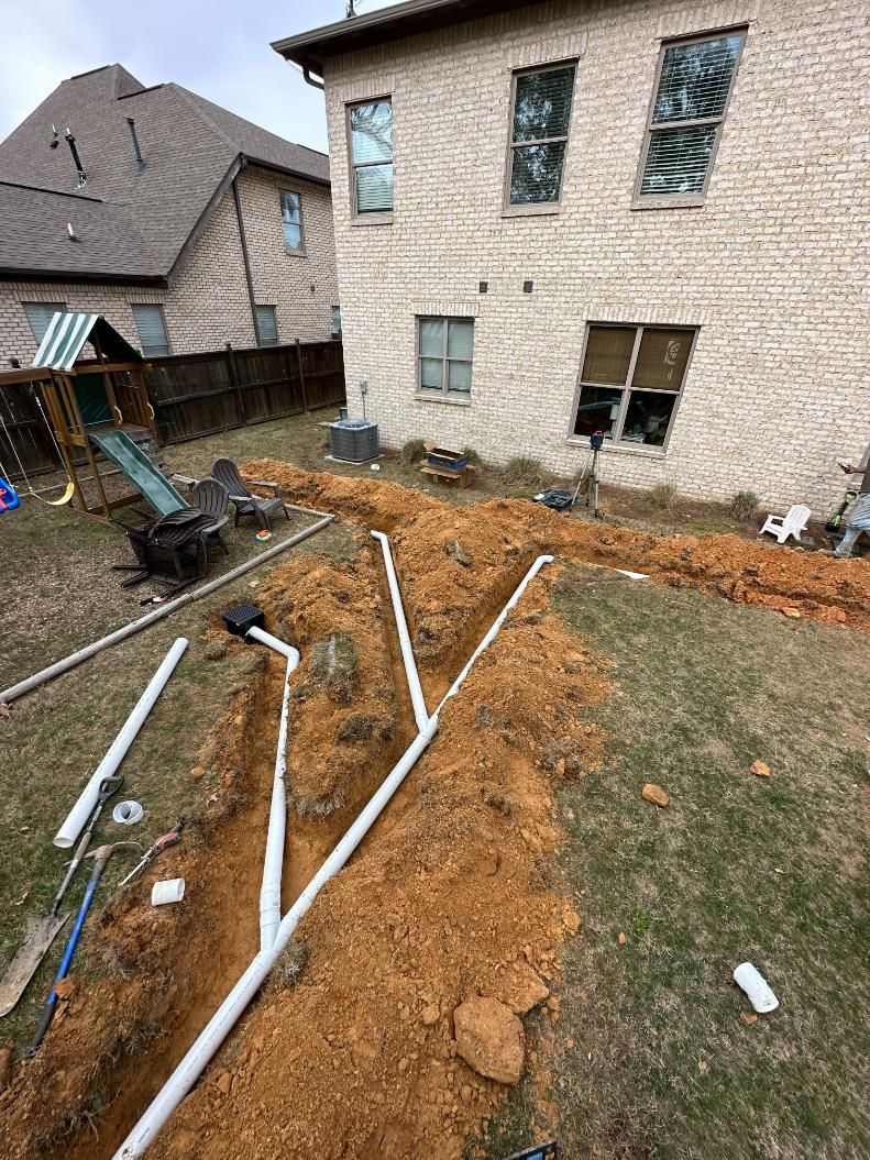 Backyard with trenches for drainage, white pipes laid in dirt, near a two-story brick house and play set.