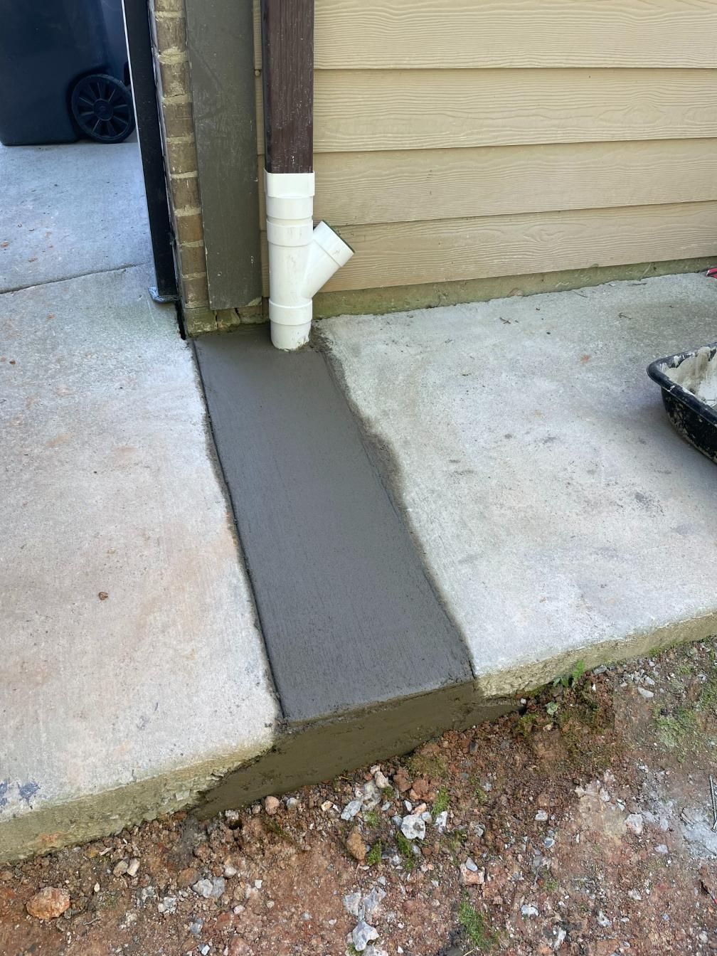 New concrete strip along a building's side, beside an existing concrete slab, with a downspout in the middle.