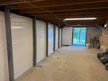 White-walled crawlspace with white plastic on the floor and support pillars, lit by overhead lights.