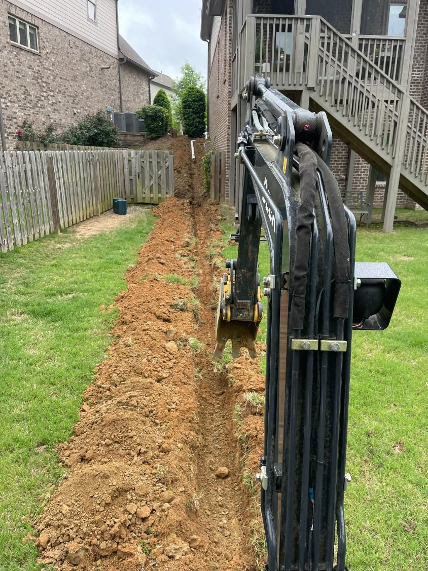 A small excavator digging a trench in a grassy backyard next to a wooden fence and house.