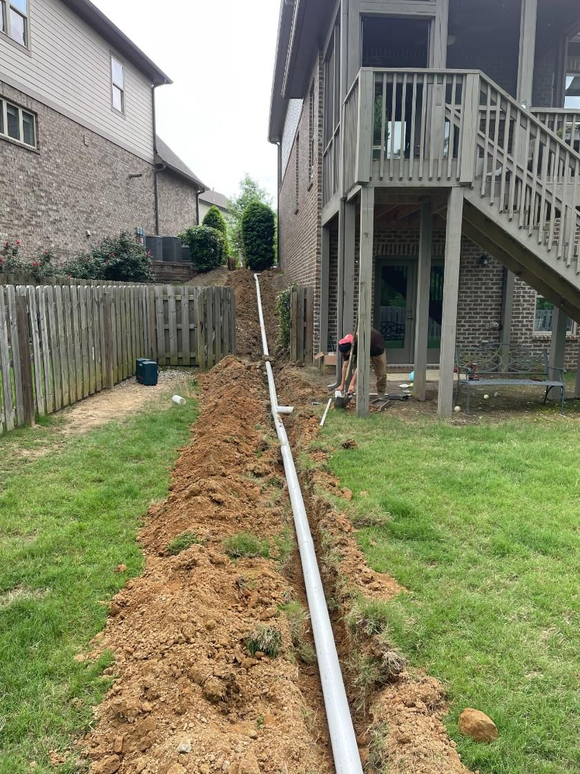 A trench dug in a backyard, with a white pipe laid inside, and two people working near the house.