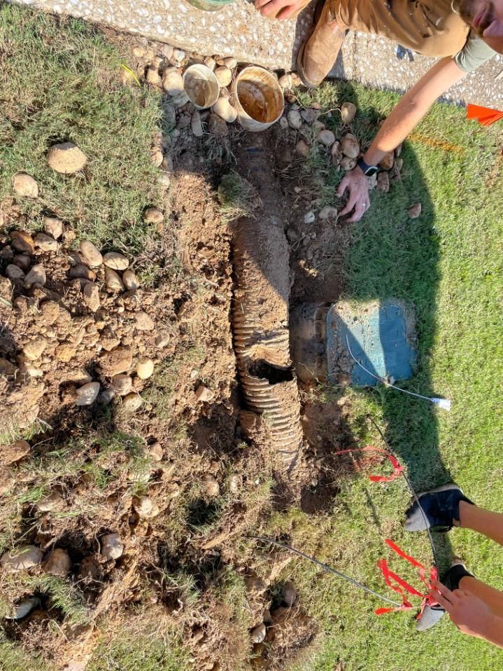 People working on a drainage system in a yard; a corrugated pipe is in the ground near grass and rocks.