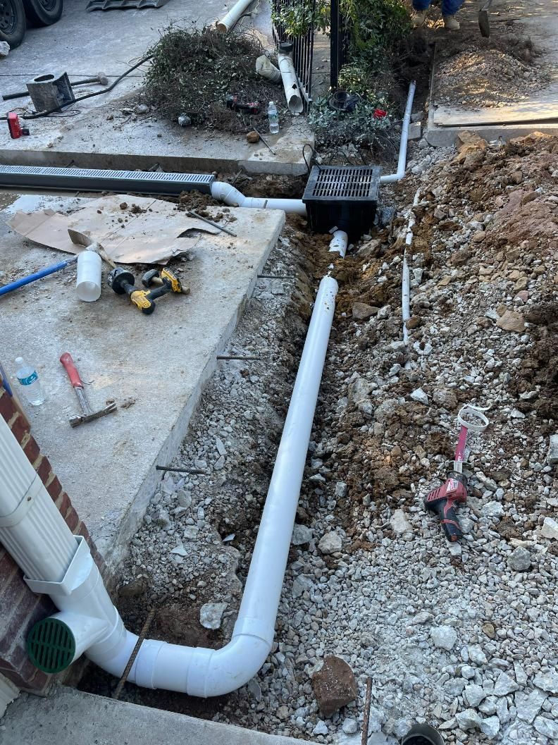 Construction site: white drain pipe installation along concrete sidewalk. Soil, tools, and a downspout visible.