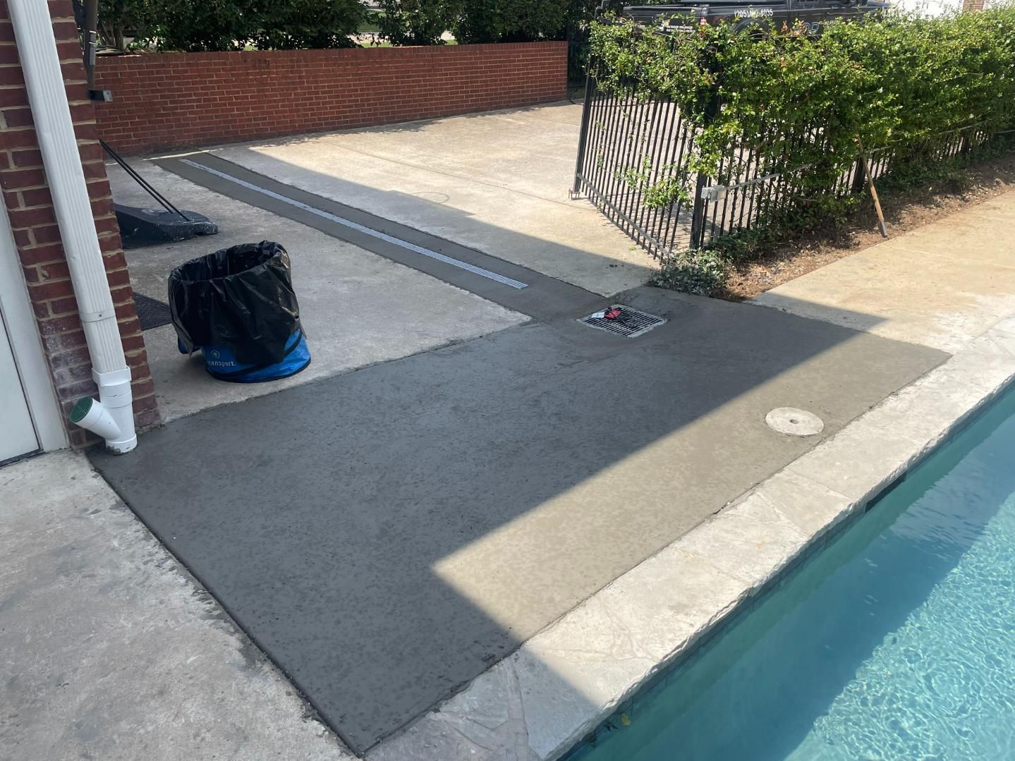 Concrete patio next to a pool; black trash bag, brick wall, and fence visible.