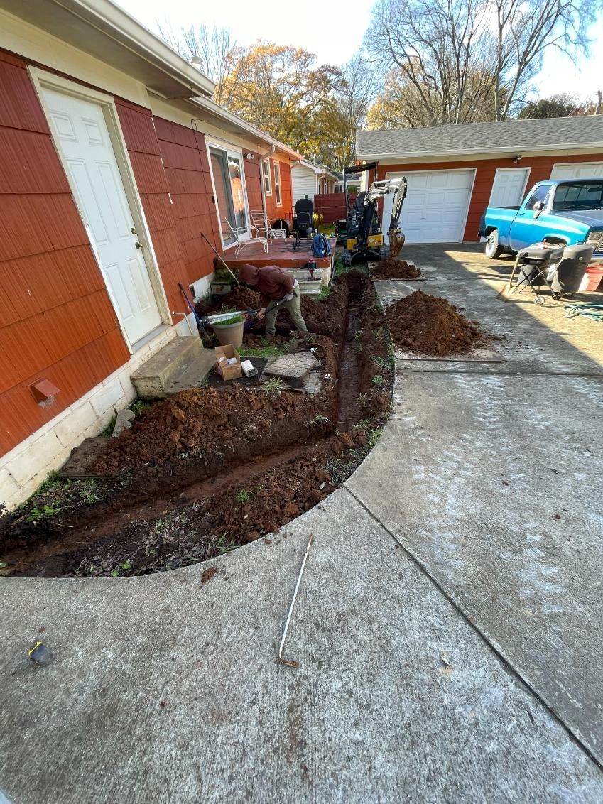 Workers digging a trench next to a red building and concrete driveway. Piles of dirt and tools visible.