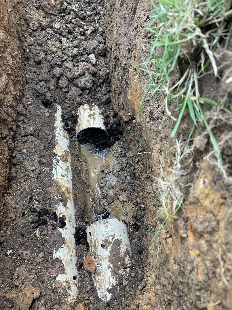 A trench in soil reveals broken white PVC pipes with black residue. Green grass borders the trench.