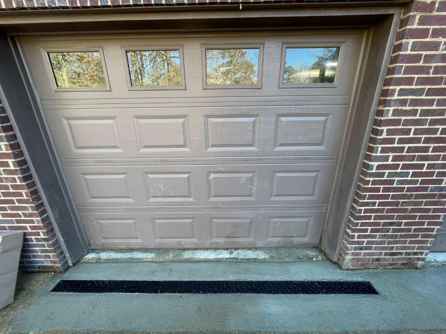 Tan garage door with windows, set in brick wall. A black drainage grate sits in front.