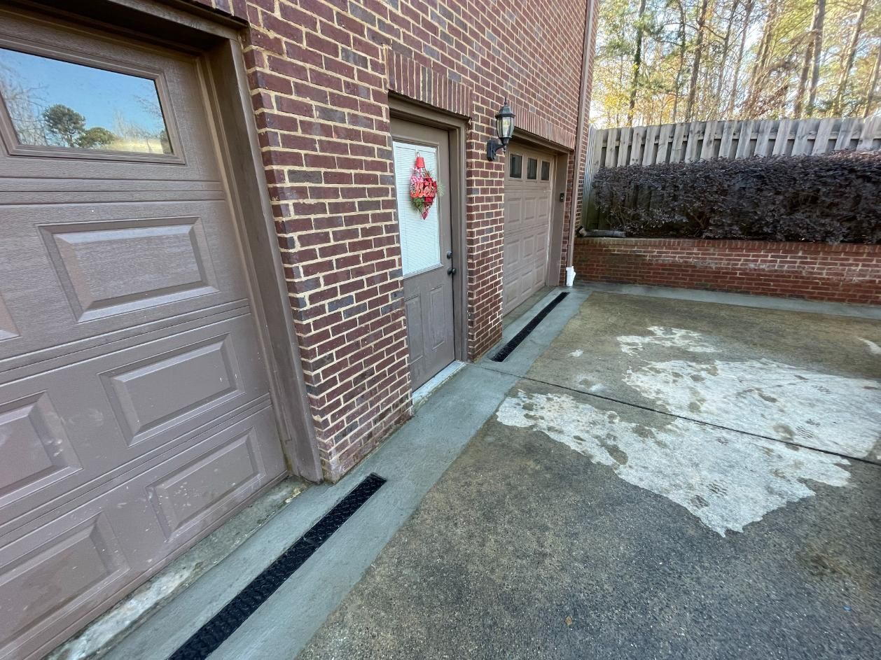 Brick garage exterior with two doors, a small door with a wreath, and a concrete driveway with a black drain.