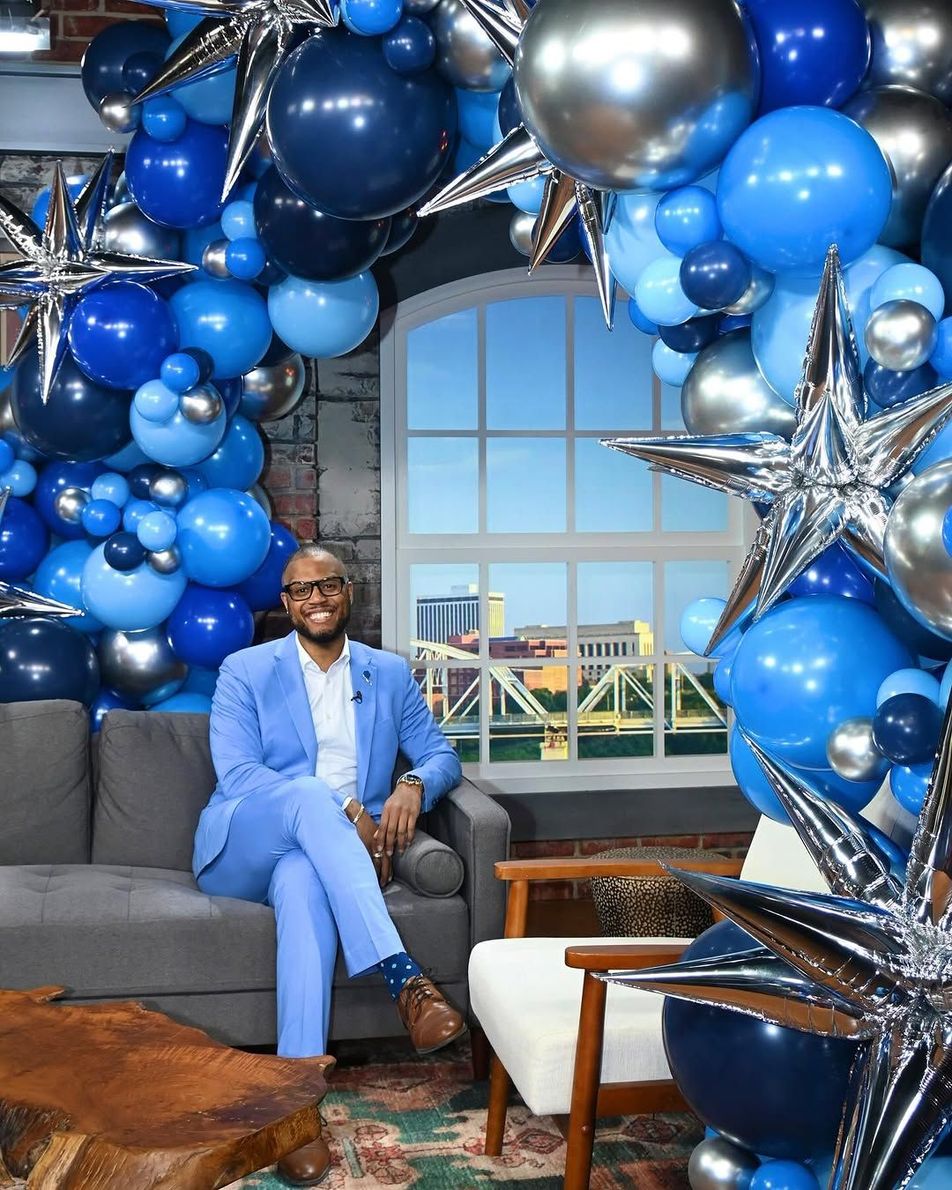 A man in a suit is sitting on a couch in front of a wall of blue and silver balloons.