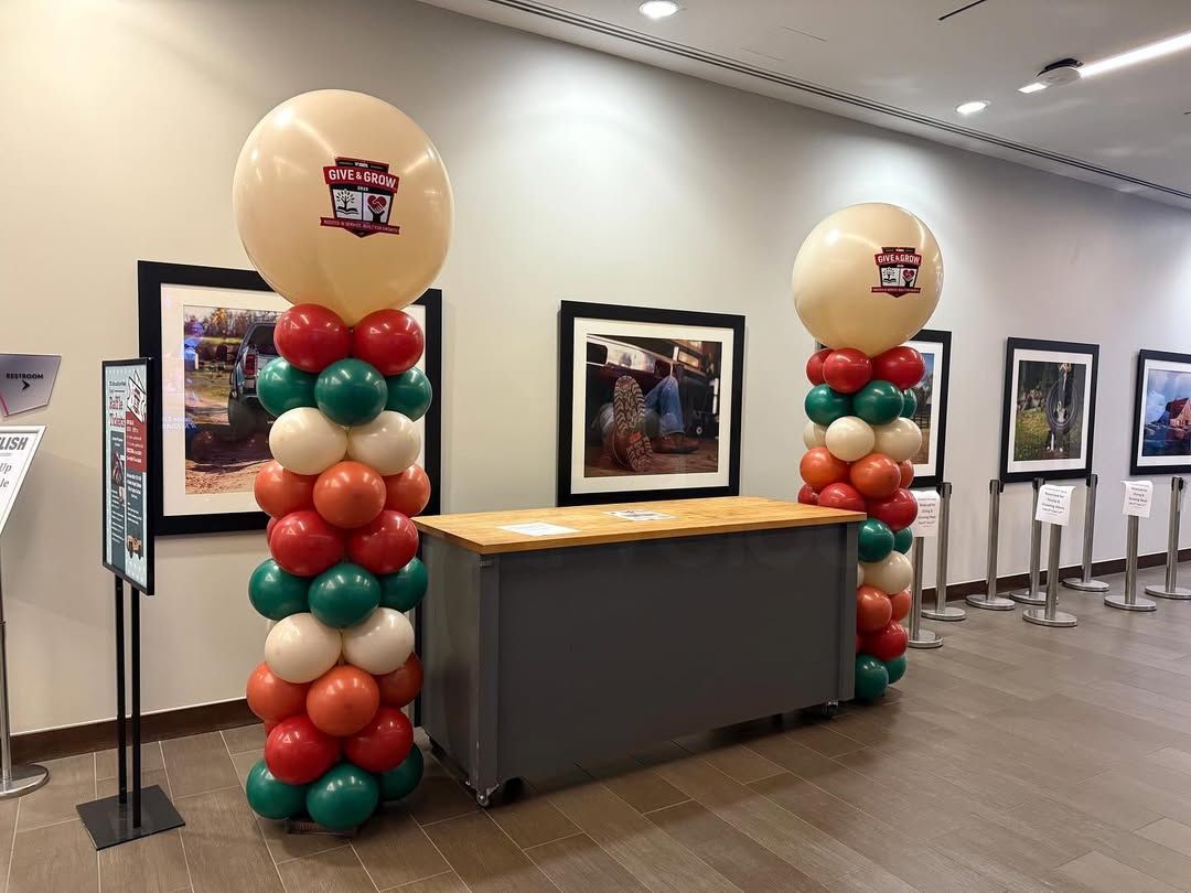 Two balloon columns with a desk in a gallery, balloons are red, green, and orange.