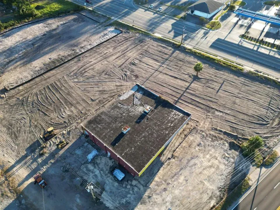 Aerial view of a small foundation site with workers, dirt lot, and nearby road and trees