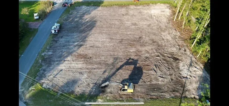 Aerial view of a cleared brown lot with a small excavator and a road along the left side.