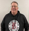 Smiling man in glasses wearing a black hoodie with a circular red-and-white logo, standing against a gray background