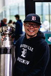 Smiling person in a black hoodie and cap stands next to a trophy in a bright indoor setting