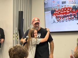Girl hugs man with microphone at indoor event, with team photo displayed on screen behind them