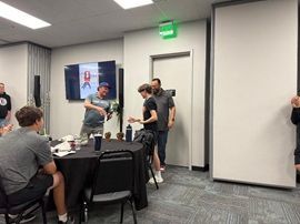 People gathered around a table in a conference room, with a screen and a green exit sign on the wall.