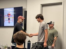 Two people shaking hands in a room, with others watching and a screen showing a hockey player.