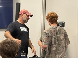Two people talking indoors near a wall, one wearing a red cap and black T-shirt, the other in a camouflage shirt.