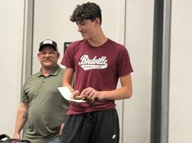 Two people standing indoors, one holding a paper and smiling, the other in a maroon shirt reading it
