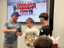 Three people standing indoors in front of a framed hockey team photo, smiling and talking.