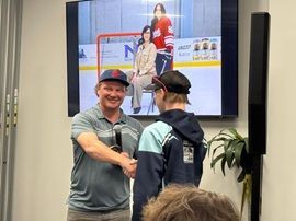 Two people shake hands in an office lobby with a hockey game photo on a wall screen behind them.