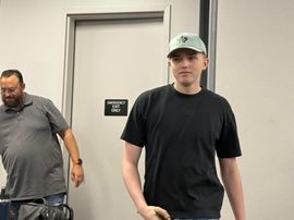 Two men standing indoors near a door, one in a black shirt and green cap, one in a gray shirt