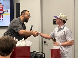 Two people shaking hands at an indoor event, with a red gift bag and a microphone nearby.