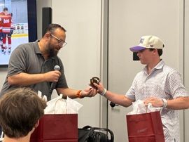 Two men exchange a small item at an event table, each holding a red gift bag.