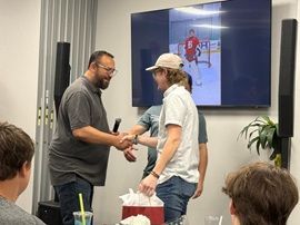 Two people shake hands during a presentation in a conference room, with a slide displayed behind them.