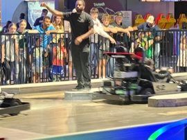 Man gesturing beside a blurred race car at an indoor event with spectators behind a barrier