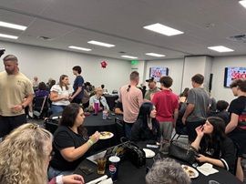Crowded community room with people chatting, eating, and watching TVs on the wall.