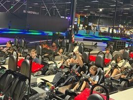 People driving go-karts at an indoor track with red and black cars under bright lights