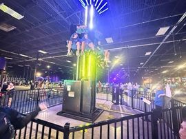 Indoor amusement ride with riders atop a glowing green-lit tower in a fenced attraction area