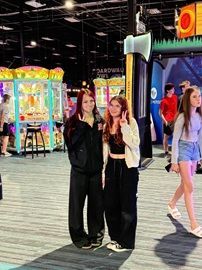 Two people posing in a brightly lit arcade, with colorful game machines in the background.