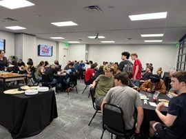People seated at tables in a community room, eating and talking during a casual gathering.