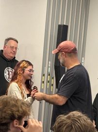 Three people at an indoor event, one handing an item to a woman while others watch.