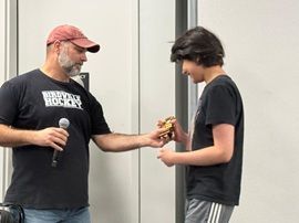 Man in red cap hands an award to a young person holding a microphone indoors