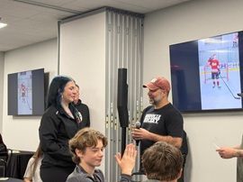 Two people speaking in a classroom, with students seated in the foreground and a screen showing a robot on the wall.