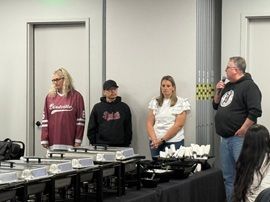 Four people stand behind a table at an indoor event, with trays and white items displayed on it.