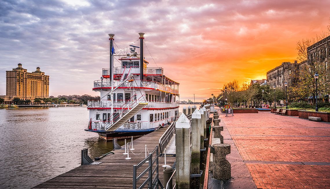 Steamboat docked at sunset on a river, with a glowing pink-orange sky and waterfront buildings nearby