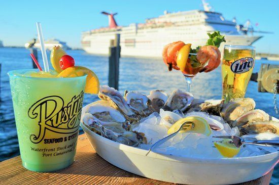 Seaside table with seafood platter and colorful cocktails, cruise ship in the background.