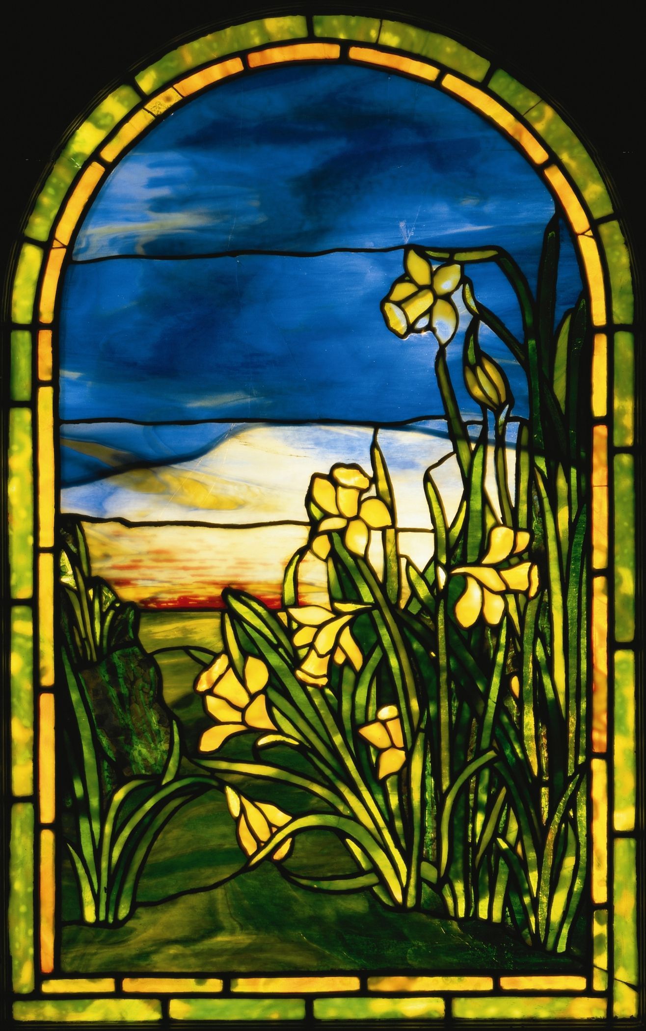 Stained-glass window with yellow flowers and tall grass against a blue sky and sunset horizon