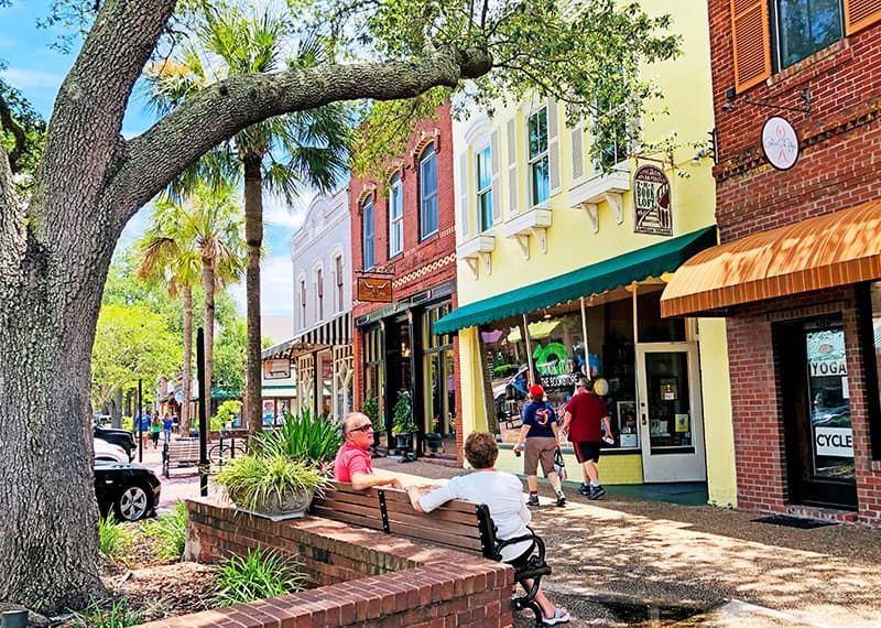 Tree-shaded sidewalk with colorful storefronts and people walking and sitting on a brick bench