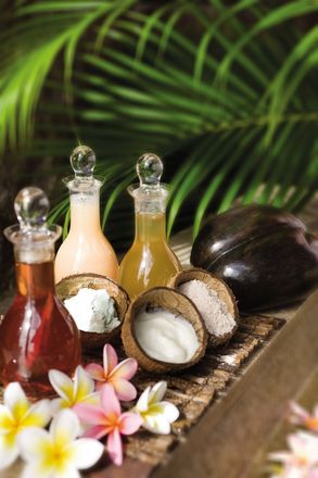 Coconut oil bottles and split coconuts with plumeria flowers on a rustic tray, against green leaves
