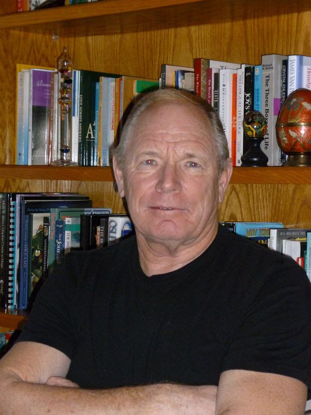 Man seated indoors before wooden bookshelves, wearing a black shirt and looking at the camera.